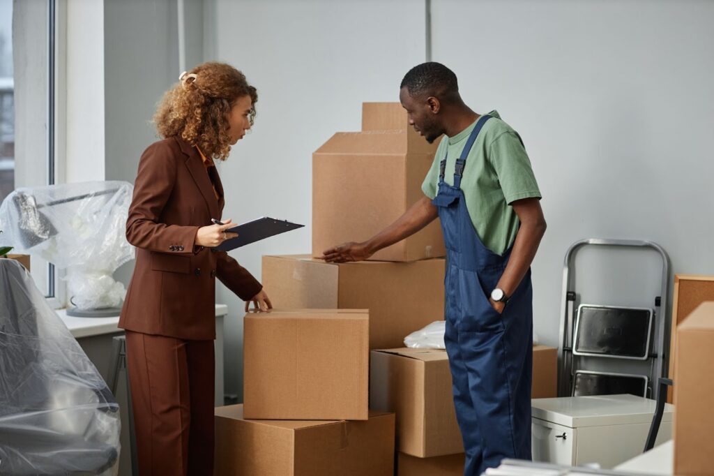 A mover assists a woman reviewing packed boxes during an office relocation.